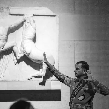 White man infront of a Greek statue, using his hands to explain the contents. 